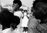 Garrett Bradley. America. 2019. Multichannel video installation. Courtesy the artist. Caption: A black-and-white image of a young Black girl in a white dress holding a reflective object that hides her face. Alt-text: Garrett Bradley. America. 2019. Multichannel video installation. Courtesy the artist. Image Description: A black-and-white image of a young Black girl stands in the background of three young Black boys in front of her, to the left, and facing left, and two Black boys in front of her, to her right, facing left, wearing grey polos and white striped tee shirts. She is facing and looking directly at the camera. She is standing in a white dress with puffed short-sleeves and socks with ruffle ankle socks poking out of the tops of her black patent leather mary janes. Her hair is pulled into two ponytails by her ears with white ribbons. She is holding an object to her chest, that is reflecting light or possibly a camera flash.