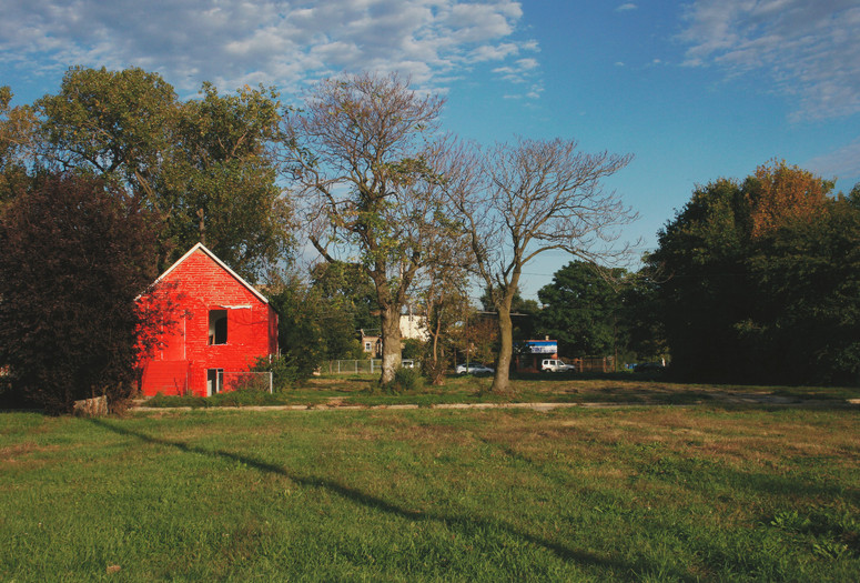 Amanda Williams. Harold's Chicken Shack, from the Color(ed) Theory Suite. 2014–2016. Pigmented inkjet print. Fund for the Twenty-First Century Alt Text: A photograph of a large grassy yard under a cloud-dotted blue sky. A concrete path winds across the yard, ending on the left at a brightly colored but worn orange-red house with no windows and a fence covered in overgrowth. Centered in the background is another wooden house, hidden behind trees and surrounded by a few parked cars. Large green and brown leafy hedges frame this residential landscape on the left and right sides, and the long shadows of trees streak across the yard.