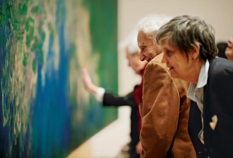 A photograph of a group of adults looking closely at a large blue and green abstract painting on the wall. Photo: Jason Brownrigg. Digital image © 2022 The Museum of Modern Art, New York