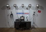 Julia Scher. Information America. 1995. Metal office desk, five 9" NTSC monitors with metal wall brackets, 13" color monitor, plastic and vinyl signage, three black-and-white surveillance cameras, removable lenses, transformers, video matrix switchers, two time-lapse recorders, Amiga A1200HD computer, Sony Watch-Cam, two media players, desk lamp, office chair, wires and cables. The Museum of Modern Art, New York. The Jill and Peter Kraus Media and Performance Acquisition Fund. © 2023 Julia Scher