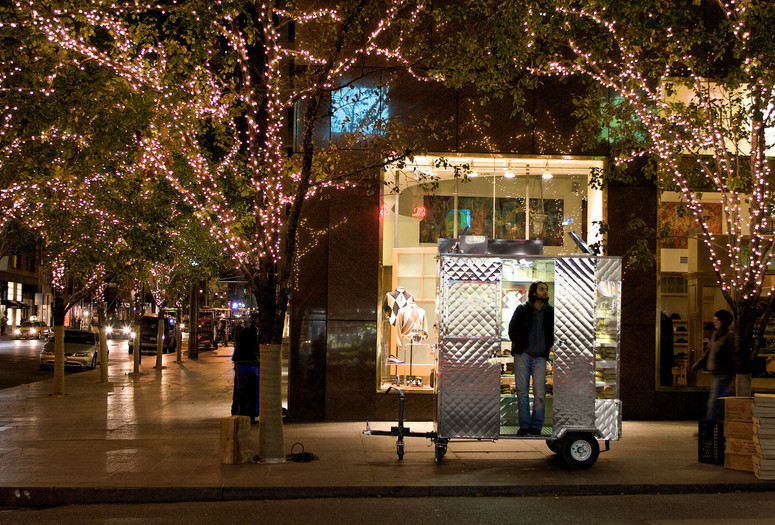 Man Push Cart. 2005. USA. Directed by Ramin Bahrani Image description: A photograph of a man standing inside his street food cart on a New York City sidewalk at night. The street is lined with twinkle light–wrapped trees, and cars and people pass by.