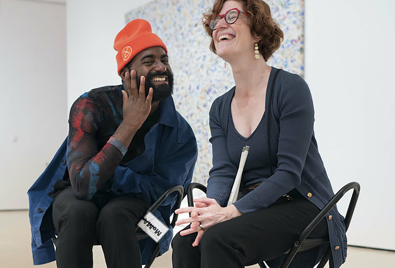 Photo: Martin Seck. ©️ 2023 The Museum of Modern Art, New York Image description: A photograph of dancer Jerron Herman and MoMA associate educator Annie Leist sitting together on black metal stools in a gallery. Jerron, a Black man wearing a neon orange beanie, holds his hand up to his face as he says something cheeky to Annie, a white woman wearing bright red glasses, who throws back her head in laughter.