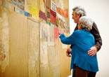 Photo: Jason Brownrigg ©️ 2024 The Museum of Modern Art, NY. Image description: Two embracing adults looking closely at the large assemblage artwork Rebus by the artist Robert Rauschenberg.
