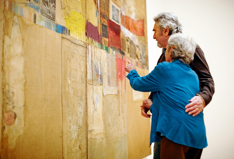 Photo: Jason Brownrigg ©️ 2024 The Museum of Modern Art, NY. Image description: Two embracing adults looking closely at the large assemblage artwork Rebus by the artist Robert Rauschenberg.