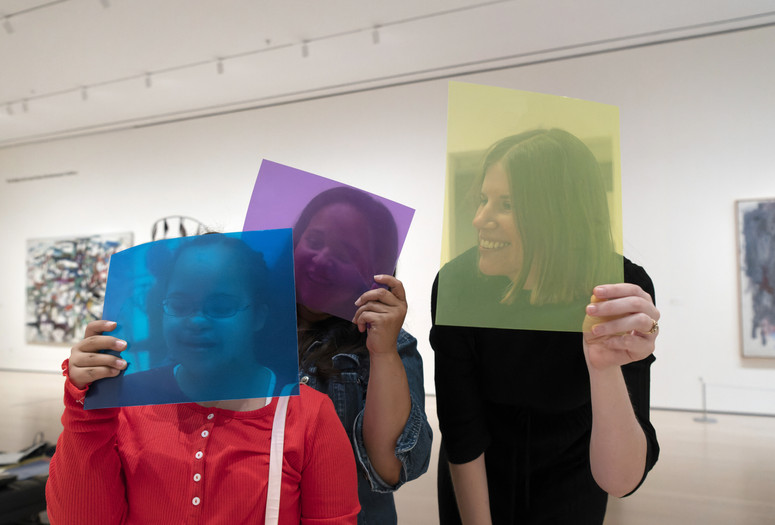 Photo: Martin Seck. © 2023 The Museum of Modern Art, New York Image description: A smiling girl in a red shirt holds a sheer blue square in front of her face alongside two smiling women holding purple and yellow squares.