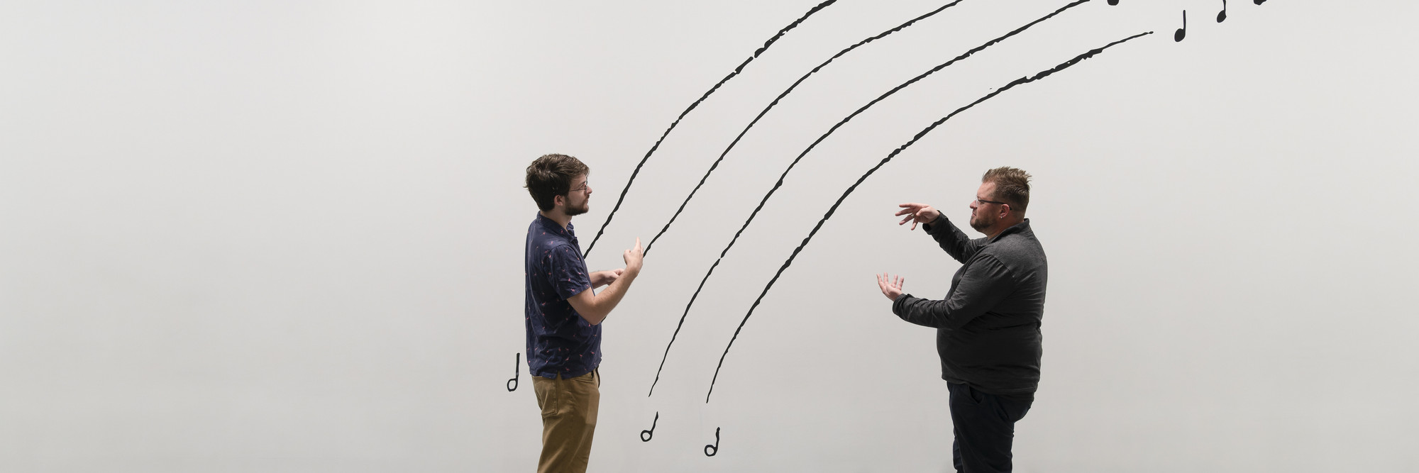 Photo: Martin Seck Image description: Two men sign to each other while standing in front of a black-and-white line drawing mural by the artist Christine Sun Kim. "The Sound of Temperature Rising" is written in capital letters at the top left corner, while the men stand in front of four arching lines that mimic music stanzas.