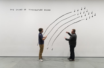 Photo: Martin Seck Image description: Two men sign to each other while standing in front of a black-and-white line drawing mural by the artist Christine Sun Kim. "The Sound of Temperature Rising" is written in capital letters at the top left corner, while the men stand in front of four arching lines that mimic music stanzas.