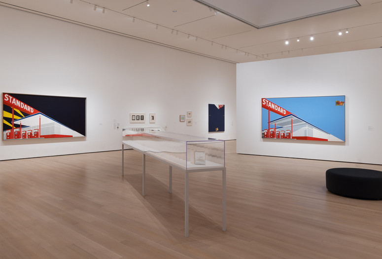Installation view of ED RUSCHA / NOW THEN, The Museum of Modern Art, New York, September 10, 2023–January 13, 2024. Photo: Jonathan Dorado. Digital image © 2023 The Museum of Modern Art, New York Image description: A gallery installation photograph with an angled glass display case in the foreground. Two large paintings of the same red Standard gas station with different coloring are in the background. The painting on the left has a dark blue sky, and the painting on the right has a bright blue sky. Smaller framed artworks line the gallery wall behind the display case.