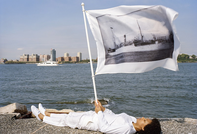 LaToya Ruby Frazier. Holding flag laying at the edge of Pier 54 and the Hudson River. On flag: US Army transport Buford, the “Soviet ark,” used to deport political radicals and other “undesirable” noncitizens from the United States to Russia in December 1919, broadside view, 1907 from Pier 54: A Human Right to Passage, 2014. © 2024 LaToya Ruby Frazier. courtesy of the artist and Gladstone gallery