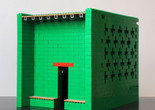 Thomas Schütte. Ackermans Tempel III (Modell 1:10) (Ackerman’s Temple III [Model 1:10]). 2011. LEGO bricks, wood, and aluminum. Collection of the artist, Düsseldorf. Photo: Luise Heuter. © 2024 Thomas Schütte/Artists Rights Society (ARS), New York/VG Bild-Kunst, Bonn Image description: A hollow cube sculpture made of green LEGO bricks resembling a temple building. A tall door opening topped with a wide red LEGO brick leads to the shadowed interior. Bricks on the right wall are stacked to create a square cross pattern. A thin piece of wood lines the rooftop, upheld by square aluminum brackets. Two wood-lined benches are built on either side of the door.