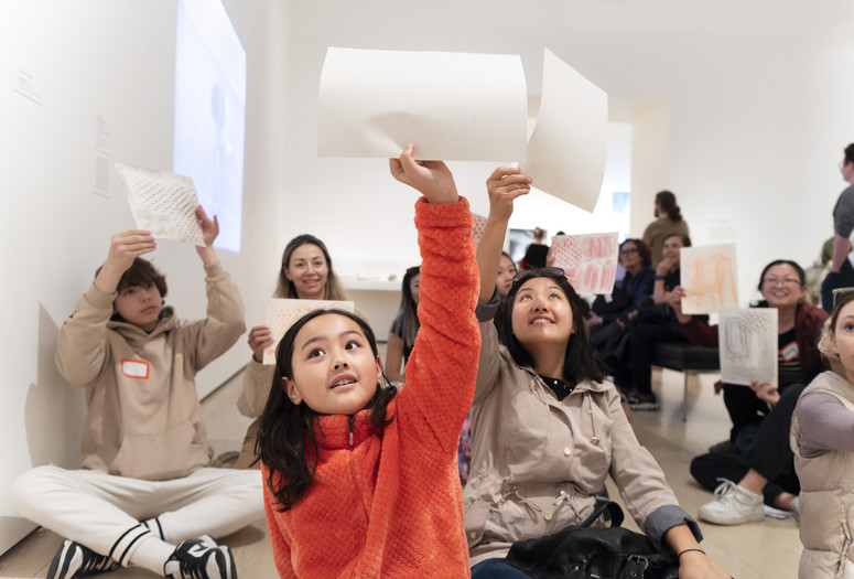 Photo: Martin SeckImage description: Children on the gallery floor hold their sheets of paper in the air.