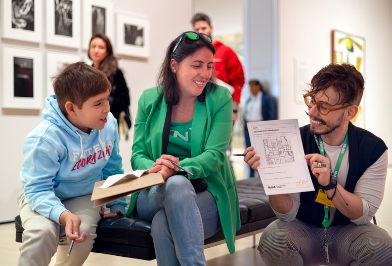 Photo: On White WallImage description: A caregiver and child look on as a smiling educator holds up a worksheet.