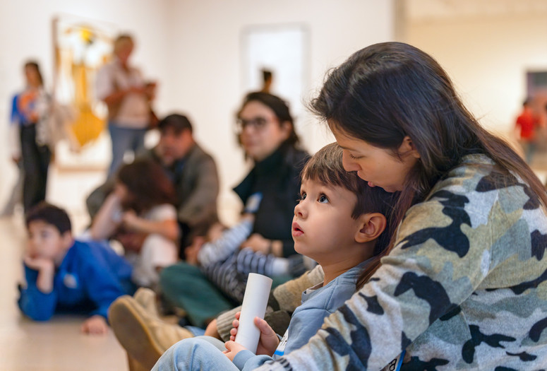 Photo: On White Wall Inc.Image description: A child sits on their caregiver’s lap on the floor of the Museum galleries while gazing up at artwork.