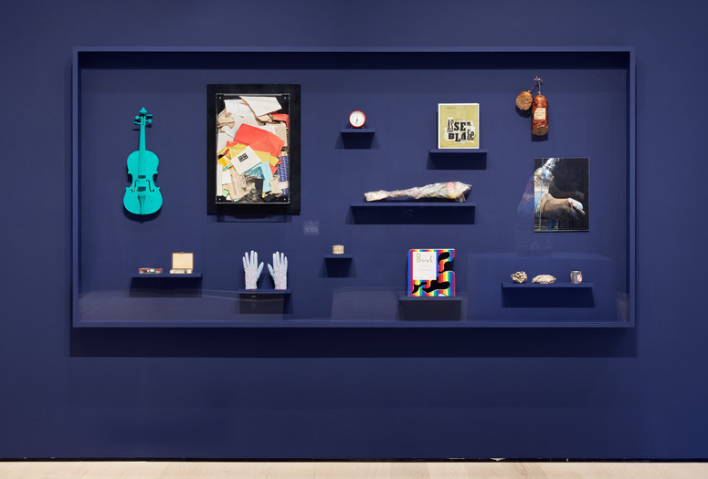 A display case with a dark blue background contains various objects, including a turquoise violin, gloves, and books. Items are arranged on black shelves, contrasting against the backdrop. Installation view, The Art of Assemblage, The Museum of Modern Art, New York, March 28, 2025–ongoing. Photographed in April 2025. Photo: Martin Parsekian. Digital Image © 2025 The Museum of Modern Art, New York
