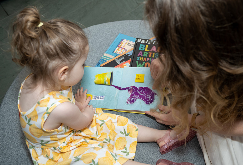 A child and an adult look at a picture book with an elephant. There are other books piled underneath, and they are sitting on a circular mat. Photo: Austin Donohue