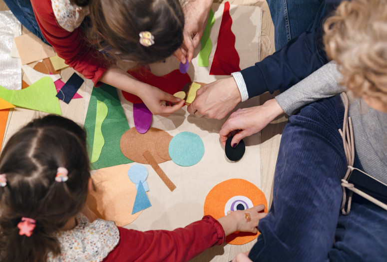 A group of four family members, photographed from above, add felt shapes of different colors to a background. Photo: On White Wall, Inc.