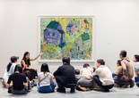 An educator leads a conversation with a group of young children and their families in front of an artwork depicting houses of different colors. Photo: On White Wall Inc.