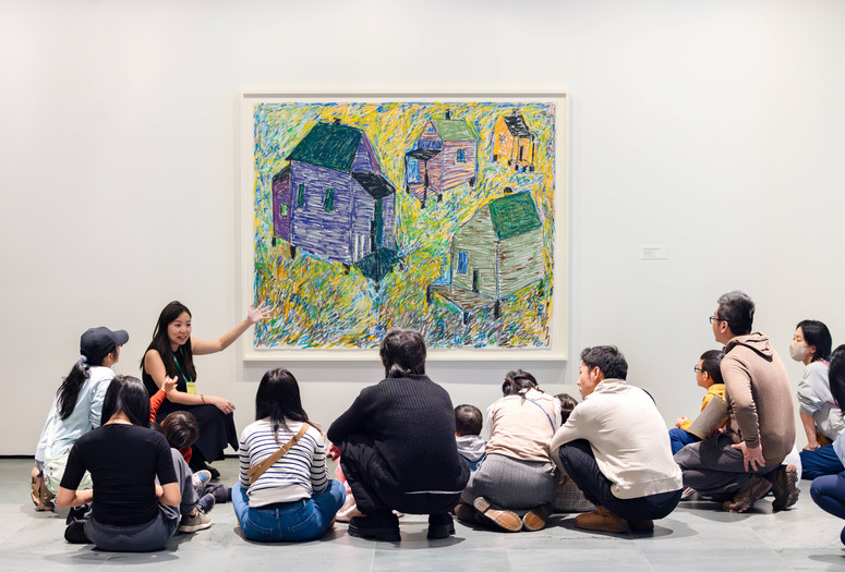 An educator leads a conversation with a group of young children and their families in front of an artwork depicting houses of different colors. Photo: On White Wall Inc.