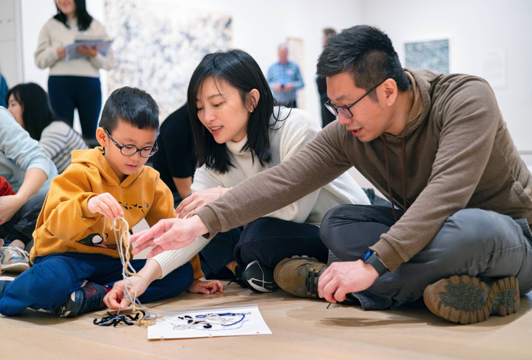 A group of people sits on the floor, engaging with a jumble of yarn on a paper. The scene is vibrant, highlighting beige and blue tones. Photo: On White Wall