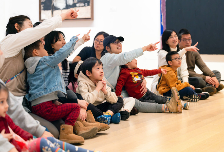 A group of adults and children sit on a wooden floor, pointing and looking in the same direction. They are wearing casual clothing in shades of blue, red, and beige. Photo: On White Wall Inc.