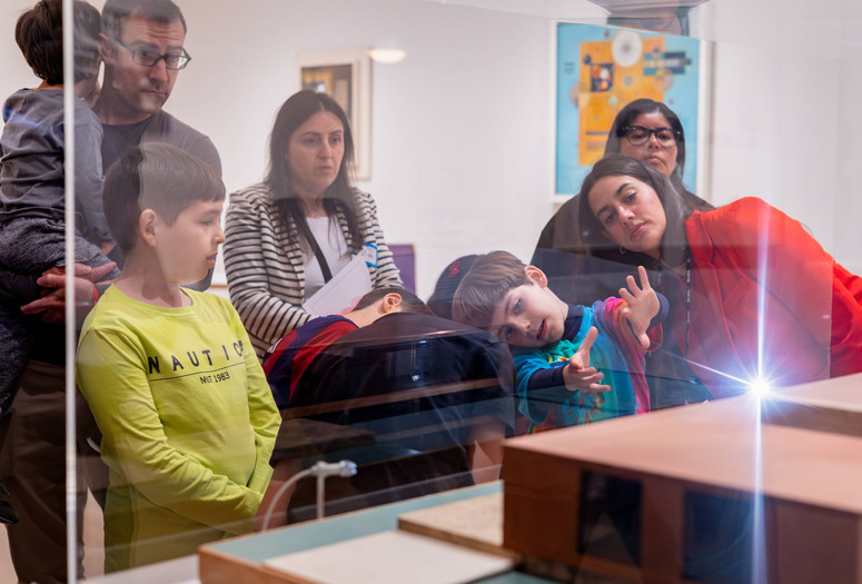 Families look at a work of art while a child and an educator tilt their heads to the left. The child holds his hands out in front of him. Photo: On White Wall, Inc.