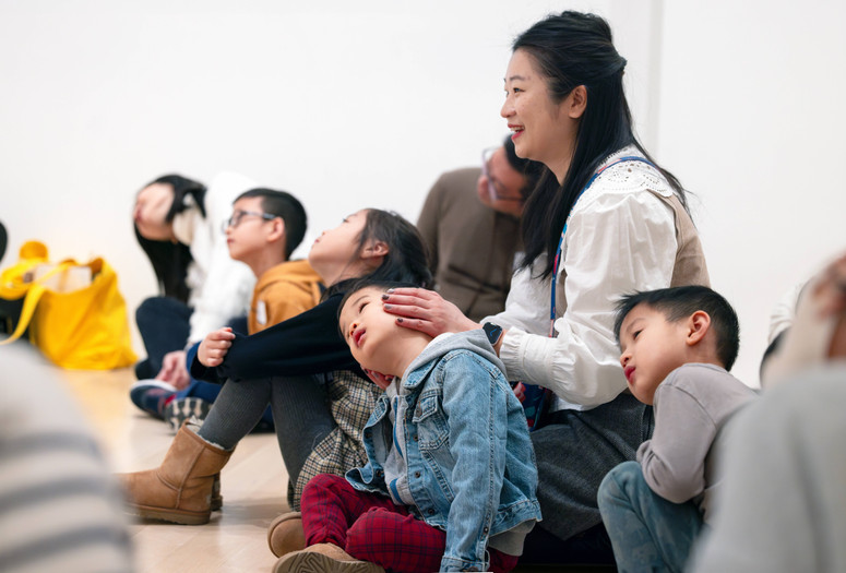 An adult holds their child’s head and turns it to the side as they sit among a group of families during a gallery talk. Photo: On White Wall, Inc.