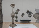Installation view of Ruth Asawa: A Retrospective, The Museum of Modern Art, New York, October 19, 2025–February 7, 2026. Photo: Jonathan Dorado Image description: A photograph of a gallery installed with several bulbous wire sculptures of various interlocking shapes hanging from the ceiling above a white base. There is a black cushioned gallery bench along the back wall.