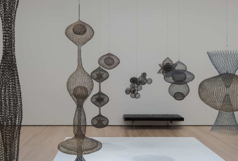 Installation view of Ruth Asawa: A Retrospective, The Museum of Modern Art, New York, October 19, 2025–February 7, 2026. Photo: Jonathan Dorado Image description: A photograph of a gallery installed with several bulbous wire sculptures of various interlocking shapes hanging from the ceiling above a white base. There is a black cushioned gallery bench along the back wall.
