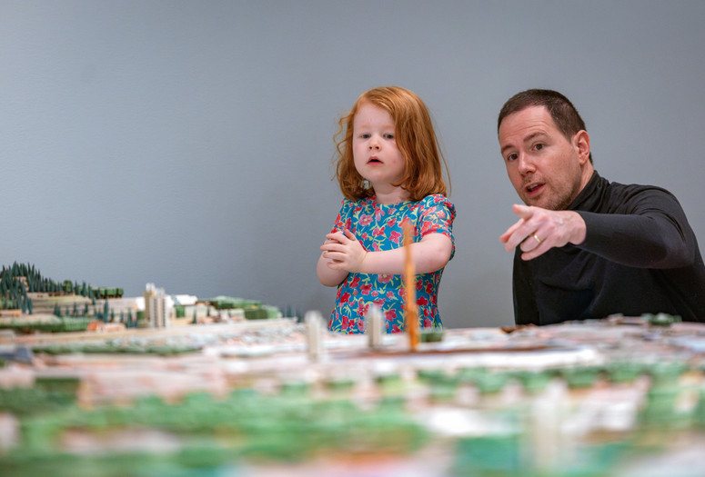 An adult points to the Broadacre City architectural model by Frank Lloyd Wright as their child looks on. Photo: On White Wall, Inc.