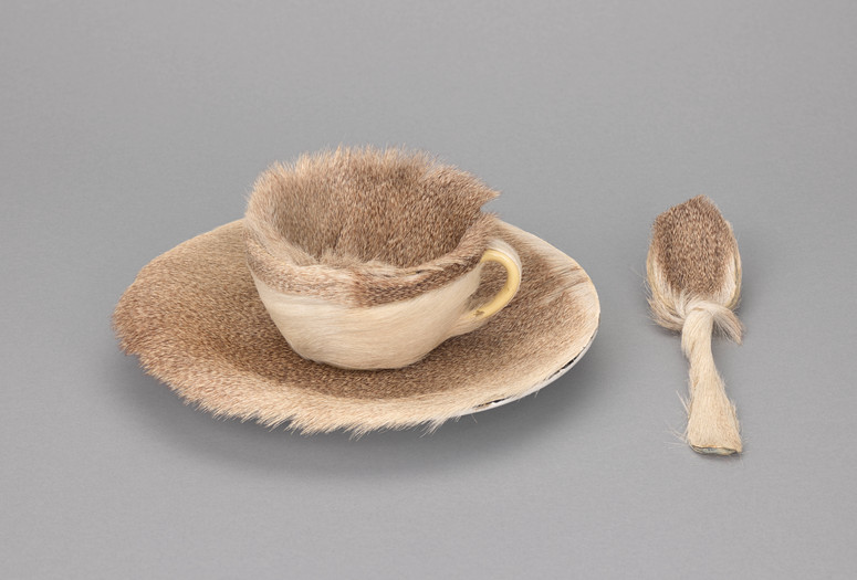 A teacup, saucer, and spoon covered in light brown fur are set against a plain grey background. The fur is thick and evenly distributed. Meret Oppenheim. Object. 1936. Fur-covered cup, saucer, and spoon. The Museum of Modern Art, New York. Purchase. © 2025 Artists Rights Society (ARS), New York/Pro Litteris, Zurich