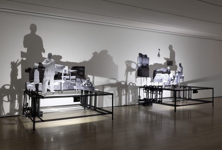 Installation view of New Photography 2025: Lines of Belonging, The Museum of Modern Art, New York, September 14, 2025–January 17, 2026. Photo: Robert Gerhardt Image description: A gallery installation featuring two platform-like steel cube sculptures against a dimly lit white wall. Black-and-white photo cutouts of a male figure standing in front of a collaged landscape on the left and a seated female figure reading in a domestic setting on the right are displayed on the structures. A bright light points towards the sculptures, casting shadows on the wall.
