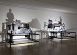 Installation view of New Photography 2025: Lines of Belonging, The Museum of Modern Art, New York, September 14, 2025–January 17, 2026. Photo: Robert Gerhardt Image description: A gallery installation featuring two platform-like steel cube sculptures against a dimly lit white wall. Black-and-white photo cutouts of a male figure standing in front of a collaged landscape on the left and a seated female figure reading in a domestic setting on the right are displayed on the structures. A bright light points towards the sculptures, casting shadows on the wall.