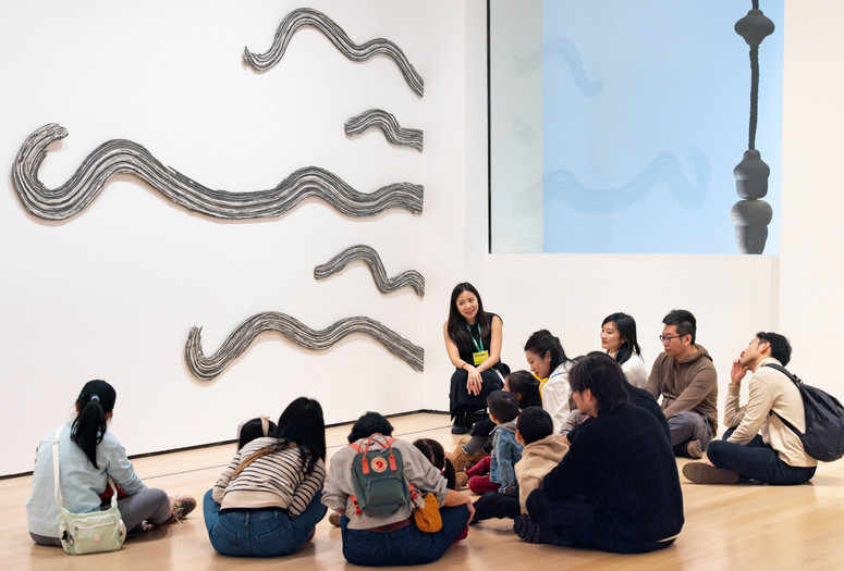 A group of children and adults and a teaching artist sit in front of an artwork. Artwork: Rosemarie Castoro. Side by Side. 1972. Modeling paste, gesso, and graphite on board, five pieces. The Museum of Modern Art, New York. Purchase. © The Estate of Rosemarie Castoro. Photo: On White Wall, Inc.