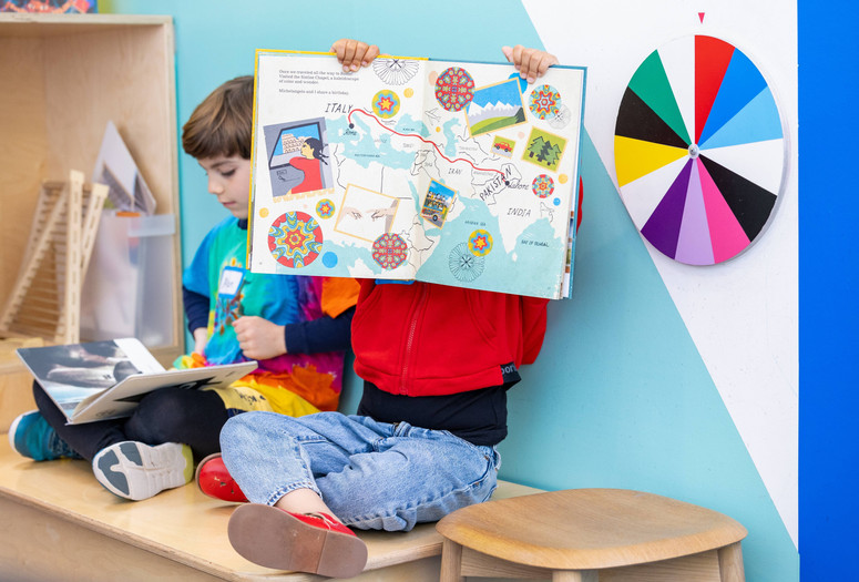 Two children sit on bench reading picture books. One holds the book on their lap, and one holds the book with the pages facing out toward the camera. Photo: On White Wall, Inc.