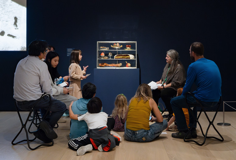 An educator leads a discussion with families in front of a sculpture of a pastry case with sculpted desserts inside. Photo: On White Wall, Inc.