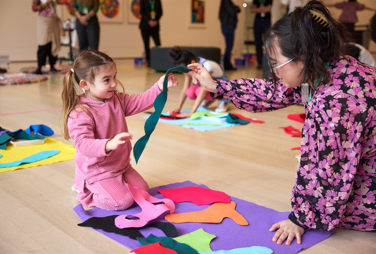 A child dressed in pink holds up a blue felt shape while she looks up at a MoMA Learning Specialist in a floral shirt. Photo: Alycia Kravitz Photography, Inc. © 2026 The Museum of Modern Art, New York