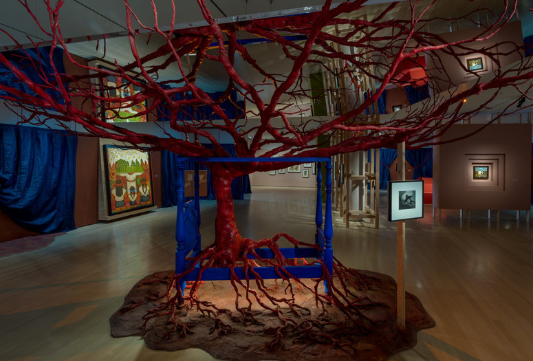 Installation view of Frida and Diego: The Last Dream, The Museum of Modern Art, New York, March 21–September 12, 2026. Photo: Jonathan Dorado. Digital image © The Museum of Modern Art, New York Image description: An installation view of a dimly lit exhibition gallery. A blood-red-colored tree in the center of the room is rooted to a mound of clay-colored earth. The tree grows up through a vibrant blue four-poster bed. Its gnarled branches reach up to cradle a large mirror hanging from the ceiling. The mirror reflects the tree within the bed below it. Paintings by the artists Frida Kahlo and Diego Rivera are spotlit on the walls in the background, framed by pulled-back blue theater curtains.