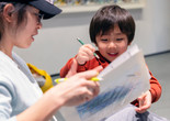 A smiling child holds a green colored pencil while his caregiver holds up two sheets of paper with blue scribbles. Photo: On White Wall, Inc.