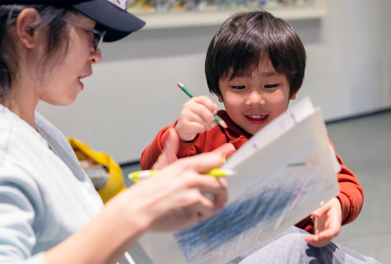 A smiling child holds a green colored pencil while his caregiver holds up two sheets of paper with blue scribbles. Photo: On White Wall, Inc.