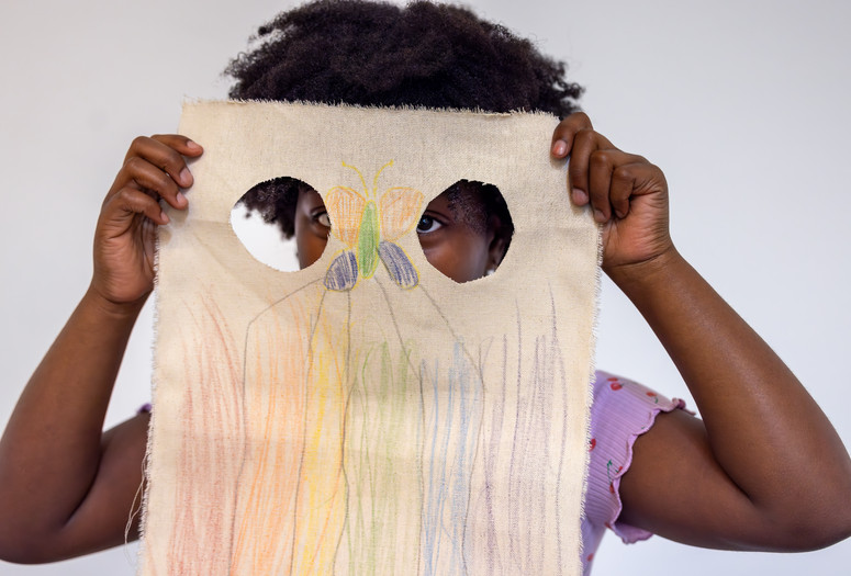 A child holds up a canvas with drawings and two holes in front of their face. Photo: On White Wall, Inc.