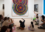 An educator leads a discussion in front of a colorful artwork by Atsuko Tanaka featuring curved lines and squiggles. Families hold up pencils in the air to trace the lines in the artwork. Photo: Martin Seck