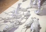 In this close-up detail of a larger installation, several aluminum foil sculptures of rats gather around a set of feline-looking paws in the top right of the image, suggesting a much larger foil animal looming just outside the frame. Though sculpted of silver foil that reflects the surroundings of a white platform and a pale gallery floor, the characters of the rats are remarkably lifelike—from their pointed noses to their tiny ears to their long thin curling tails—with the potential to scurry away at any moment. Dean Millien. The Cats and the Rats. 2026. Aluminum foil. Courtesy the artist. Photo: John Kim