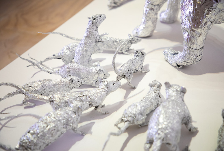 In this close-up detail of a larger installation, several aluminum foil sculptures of rats gather around a set of feline-looking paws in the top right of the image, suggesting a much larger foil animal looming just outside the frame. Though sculpted of silver foil that reflects the surroundings of a white platform and a pale gallery floor, the characters of the rats are remarkably lifelike—from their pointed noses to their tiny ears to their long thin curling tails—with the potential to scurry away at any moment. Dean Millien. The Cats and the Rats. 2026. Aluminum foil. Courtesy the artist. Photo: John Kim