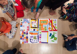 Seven children sit on the floor of a gallery around a large paper with colorful tape and tissue paper affixed to it. Photo: On White Wall, Inc.
