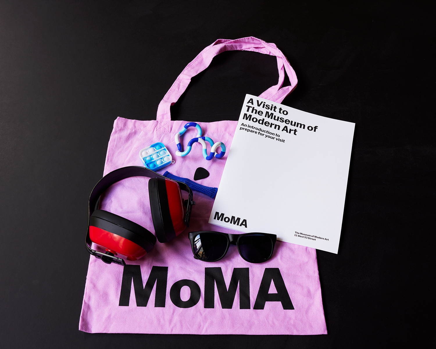 Photo: Jason LindbergImage description: Bright pink MoMA tote covered by sensory kit materials, including the social guide white booklet, over-ear noise cancelling headphones, black sunglasses, a black guitar pick, three blue fidget toys.