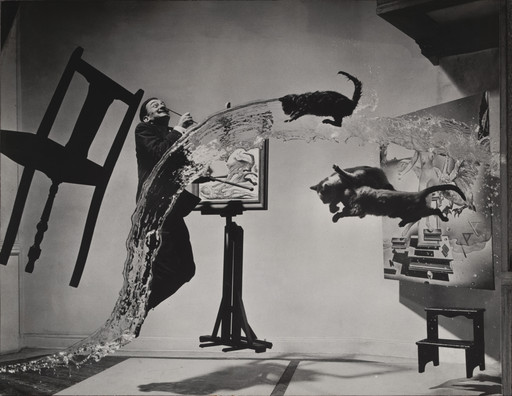 A person stands next to an easel, which holds a painting, while water and three cats are suspended mid-air. Surrounding them is a floating chair and a small table against a gray backdrop. This is an AI generated description. To give feedback, email ai@moma.org.