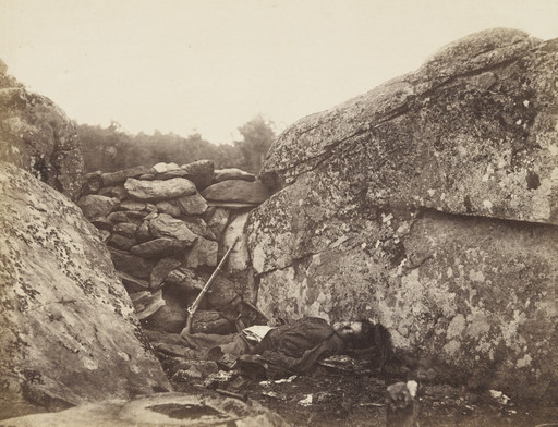A person lies on the ground between large, lichen-covered rocks with a rifle resting nearby. In the background, there is a low wall made of stacked stones and a treeline. This is an AI generated description. To give feedback, email ai@moma.org.