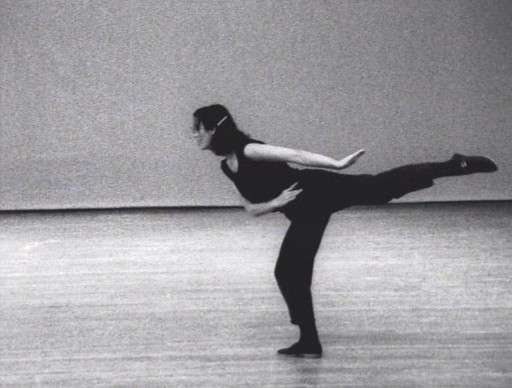 A person dressed in black is on stage, balancing on one leg with the other leg extended straight behind them, and their body leaning forward. The background and floor are gray. This is an AI generated description. To give feedback, email ai@moma.org.