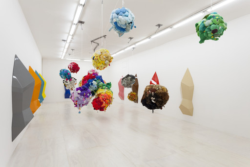 Sculptures of colorful, clustered shapes hang from the ceiling in a gallery. Bright hues of red, blue, yellow, and green contrast with geometric wall forms. This is an AI generated description. To give feedback, email ai@moma.org.