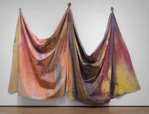 Colorful fabric draped on a wall, featuring a mix of pink, purple, yellow, and orange hues. The textile art creates a cascading, fluid appearance. This is an AI generated description. To give feedback, email ai@moma.org.